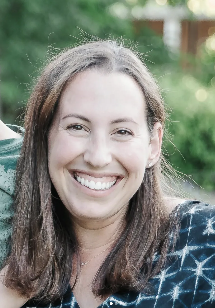Smiling woman with straight brown hair, wearing a dark patterned top, standing outdoors with greenery in the background.